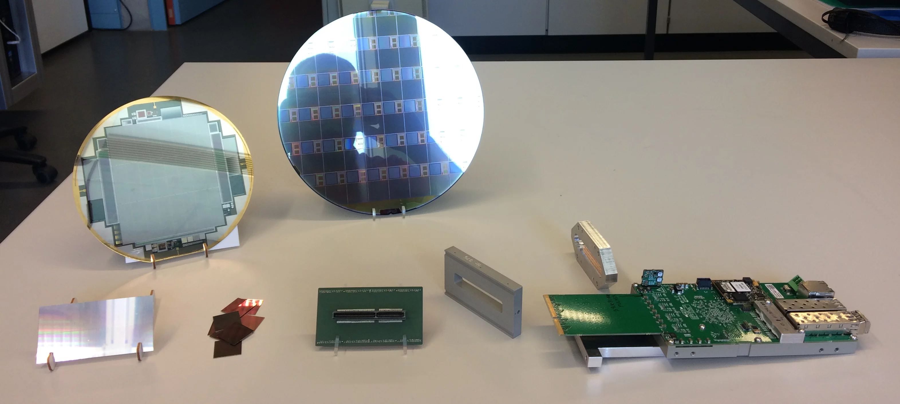 Components of a JUNGFRAU module. From left to right, in the first row: Sensor, ASICs, High Density Interconnect, aluminum holder, readout board. In the background you can see the sensor wafer and the ASIC wafers.