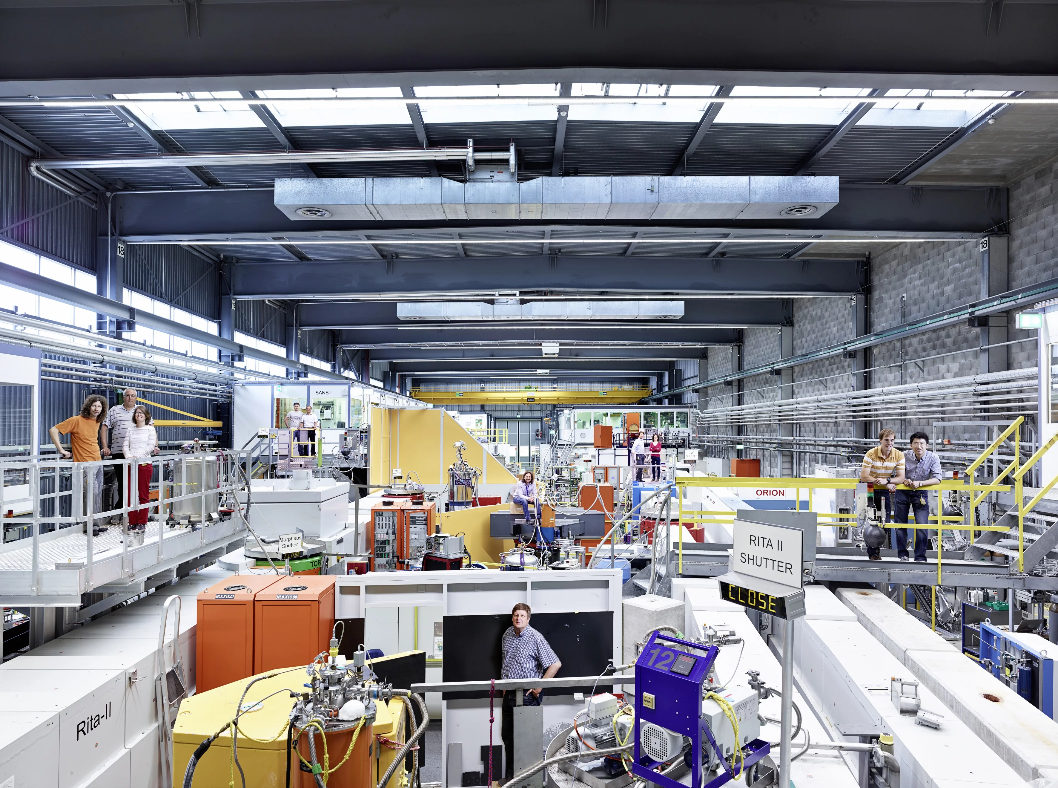 View into a part of the hall where experiments with neutrons are carried out at the Paul Scherrer Institute PSI. The photo is taken from the viewpoint of the neutron source. You can see some of the individual experimental setups, which are distributed throughout the hall. Inside the concrete channels are neutron guides, which lead neutrons to the experiment stations at the end of the hall. (Photo: Scanderbeg Sauer Photography)