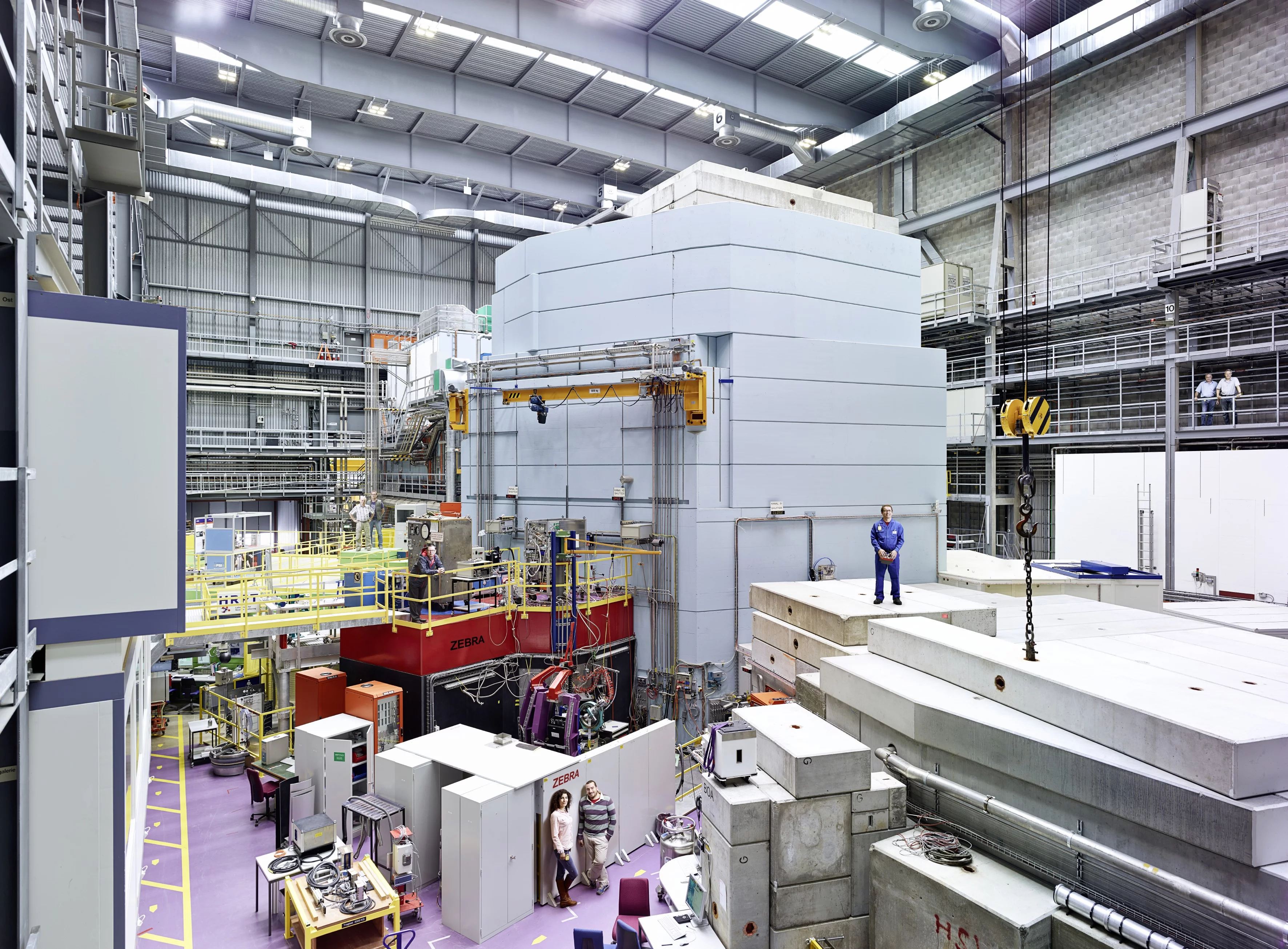 Part of the experiment hall at the Swiss Spallation Neutron Source SINQ of the Paul Scherrer Institute PSI. Plainly visible in the middle of the picture is a light-blue steel and concrete structure: the neutron source. Inside it there is a water-filled container and within that, in turn, sits a block of metal from which a beam of fast protons knocks out neutrons for the experiments. Experiment stations are arranged around this neutron source. (Photo: Scanderbeg Sauer Photography)