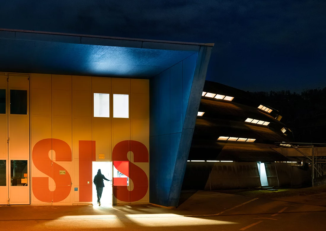 Auch die Synchrotron Lichtquelle Schweiz SLS ist mit ihren vielen Messplätzen rund um die Uhr in Betrieb. Von oben betrachtet wirkt das kreisrunde, Ufo-ähnliche Gebäude in der Dämmerung eher unscheinbar (siehe die SLS auf dem Luftbild des PSI, links oben). Der beleuchtete Eingang vermittelt dagegen einen Eindruck von der Grösse der Anlage.