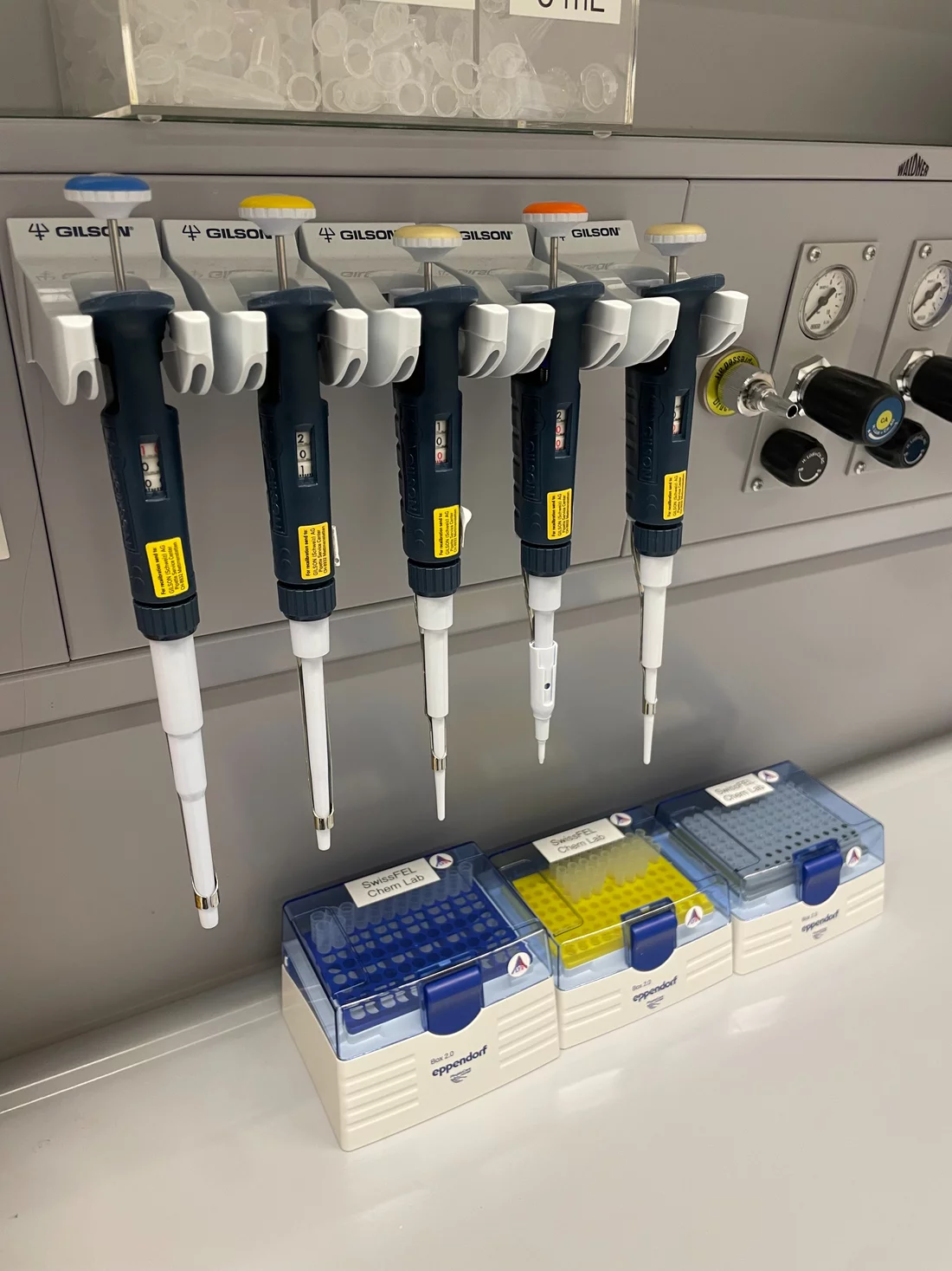 5 blue and white pipettes hanging in their holders above 3 boxes of pipette tips