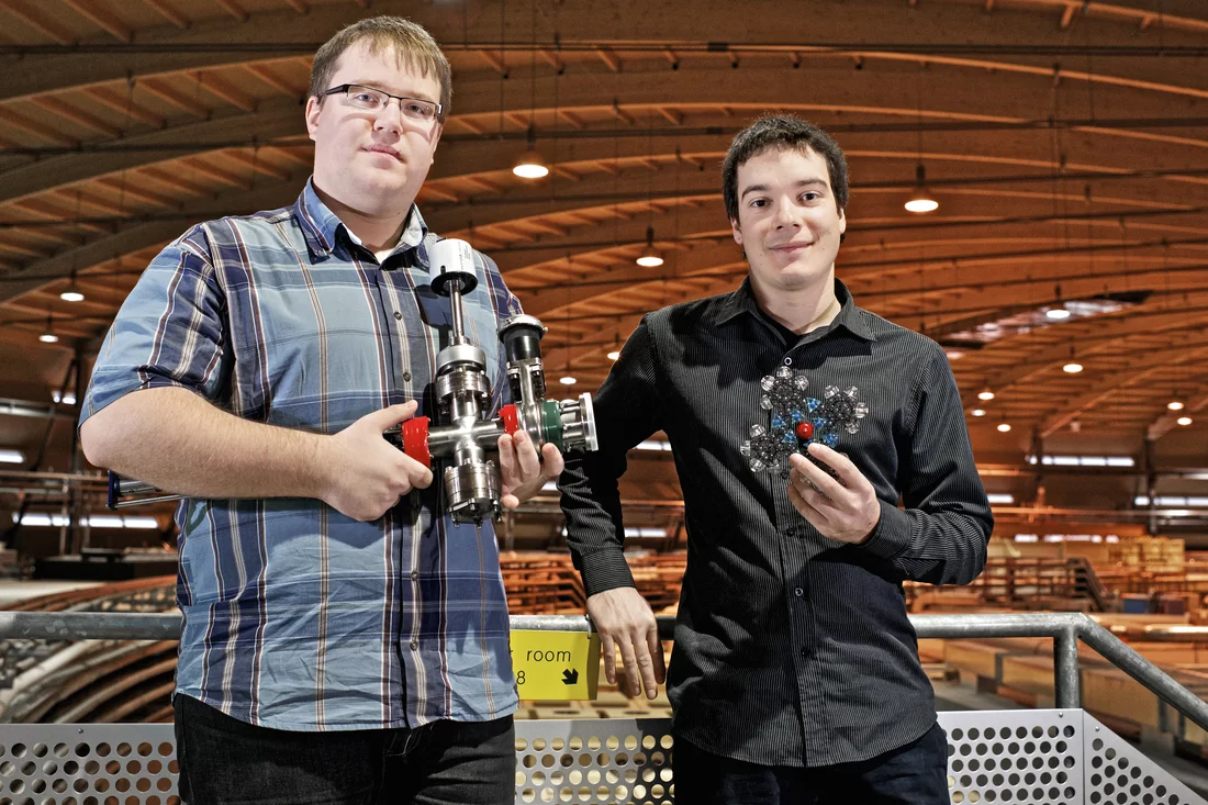 Die PSI-Forscher Jan Nowakowski (links) und Christian Wäckerlin in der Halle der Synchrotron Lichtquelle Schweiz SLS. Nowakowski hält einen „Vakuum-Koffer“, in dem die Proben für die Untersuchung zur SLS transportiert werden, Wäckerlin das Modell eines der verwendeten Moleküle. (Foto: Paul Scherrer Institut/Markus Fischer)