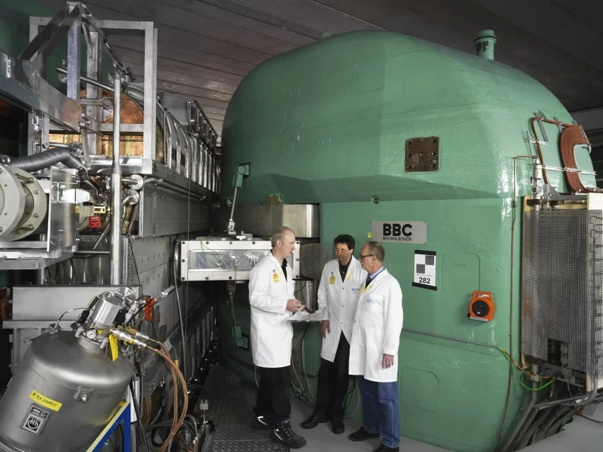 Three lofty men beside an even bigger magnet of the ring cyclotron. In the middle is Joachim Grillenberger, and on his right his predecessor Stefan Adam. On the left in the photo is one of the four cavities which accelerate the protons on their circular trajectory. 