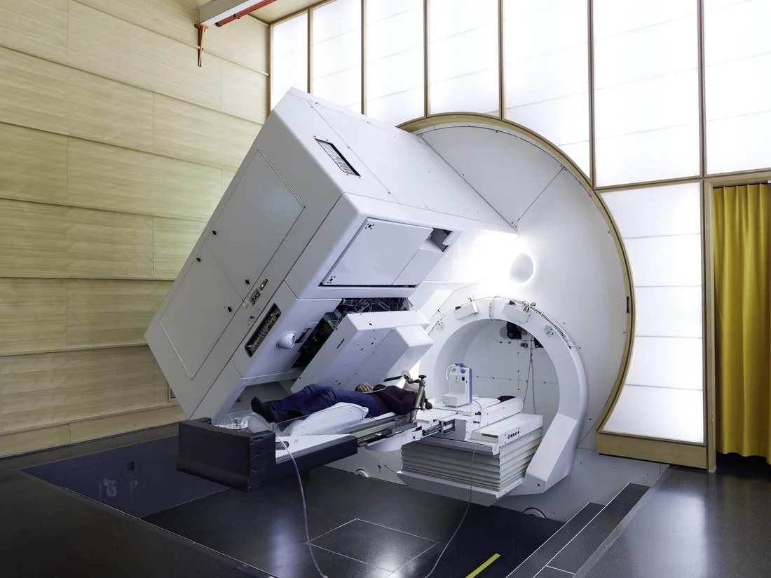 Patient irradiation at Gantry 1 of the Paul Scherrer Institute. At this facility on 25.11.1996, the first patient worldwide was treated with the spot-scanning technique, which enables particularly efficient and conservative proton irradiation of tumours. The proton beam comes out of the black slit that can be seen on the left beside the patient’s head. Behind the slit are the absorber plates that make it possible to precisely determine the beam’s penetration depth. The entire irradiation unit can rotate ar…