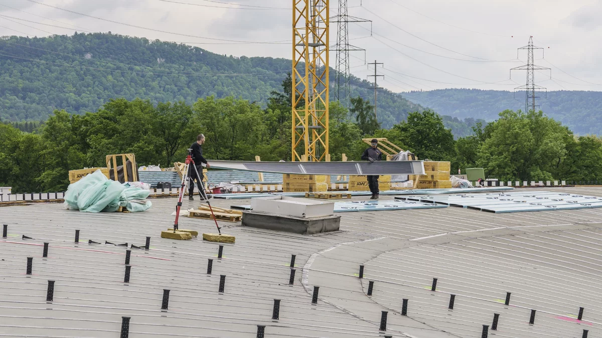 View over the main roof of the SLS. In the foreground you can see the black spacers already installed at regular intervals. In the background, no spacers have yet been installed. Two workers are walking across the roof, transporting an aluminium profile several metres long.