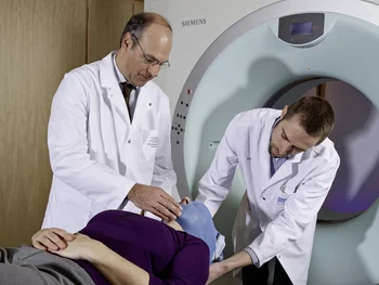 Damien Charles Weber, directeur du Centre de protonthérapie au PSI, prépare la patiente avec un assistant médicotechnique pour un examen au moyen de la machine de tomographie numérique. Cette machine se trouve dans la même salle que l'installation de traitement Gantry 2. Elle permet de contrôler la bonne position du patient juste avant le traitement au moyen d'un examen par tomographie numérique. (Photo: Scanderbeg Sauer Photography)