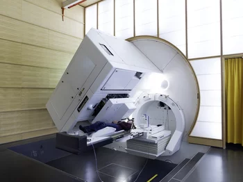 Irradiation d'un patient à la Gantry 1 de l'Institut Paul Scherrer. Le 25 novembre 1996, en première mondiale, un patient était traité au moyen d'un nouveau procédé, la technique Spot Scan, qui permet une irradiation des tumeurs avec des protons particulièrement efficace, tout en ménageant les tissus sains. Le faisceau de protons jaillit par la fente noire que l'on aperçoit à gauche, à la côté de la tête de la patiente. Derrière la fente se trouvent les plaques absorbantes qui permettent de déterminer préc…