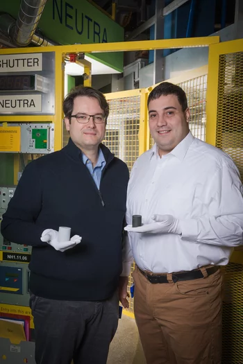 Die beiden PSI-Forscher David Mannes (links) und Christian Grünzweig vor der Experimentieranlage Neutra. Hier haben sie die Neutronenbilder von Keramikbauteilen des Technologieunternehmens ABB angefertigt. Beide Forscher halten je einen der verschiedenen Ausführungen dieser Keramik-Varistoren in der Hand: der linke, helle, ist noch nicht ausgebrannt, der rechte, dunkle, ist es bereits. (Foto: Paul Scherrer Institut/Mahir Dzambegovic)
